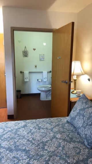 View from a bedroom into an adjacent bathroom. The bedroom has a bed with a blue and white patterned bedspread and a bedside table with a lamp and a cup. The bathroom has a toilet, a grab bar, two towels hanging on the wall, and some decorative wall hangings.