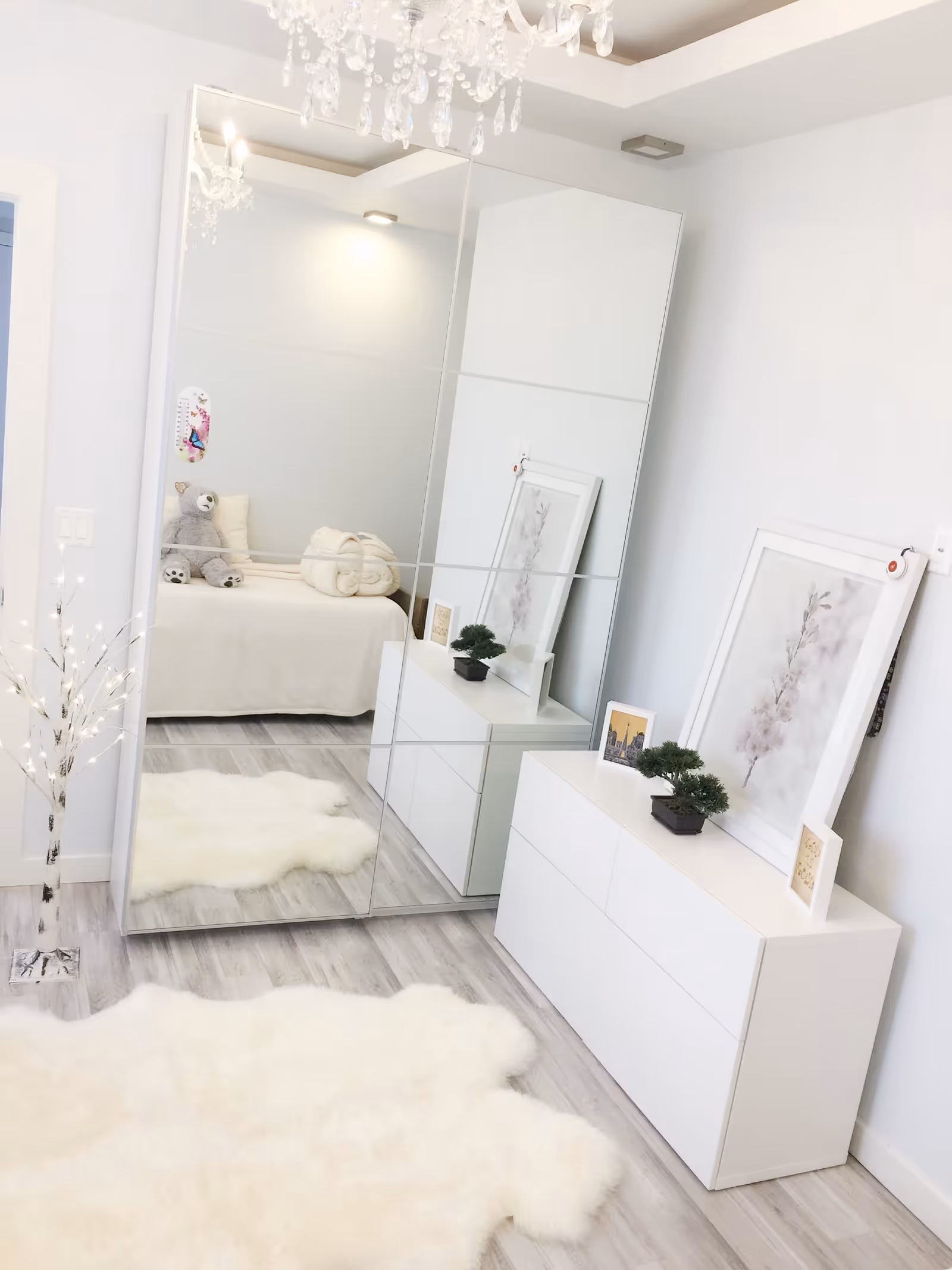 A bright and clean bedroom with light wood flooring and white walls. A large mirrored wardrobe reflects a bed with a beige blanket, a teddy bear, and a rolled towel. A white dresser with framed pictures and small potted plants sits against the wall. A fluffy white rug covers part of the floor, and a decorative lighted tree stands near the wardrobe. A chandelier hangs from the ceiling.