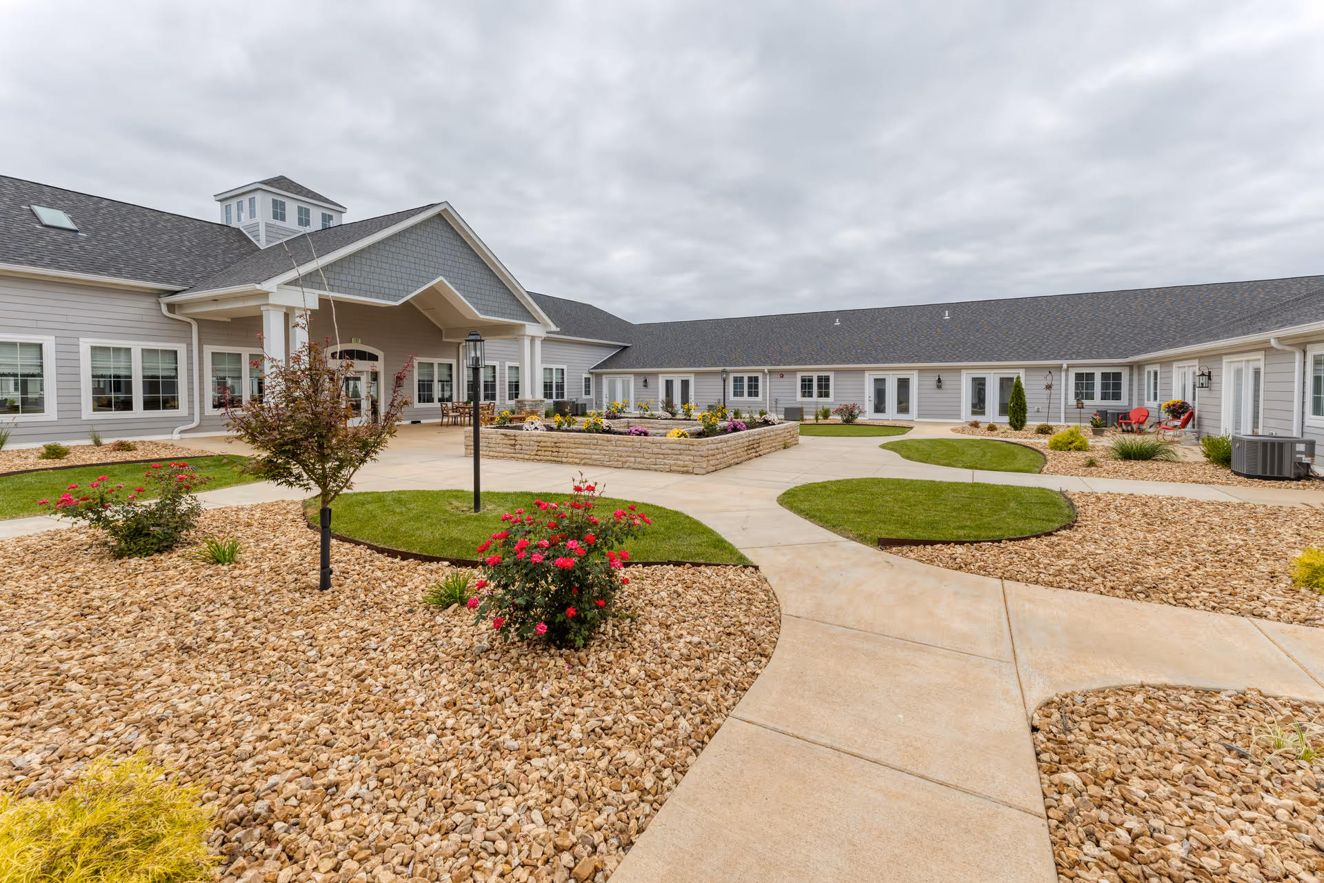 Outdoor courtyard area of Cedarhurst Senior Living of West Plains featuring a paved walkway, landscaped garden beds with rocks, small trees, and flowering plants, surrounded by a single-story building with multiple windows and doors under a cloudy sky.