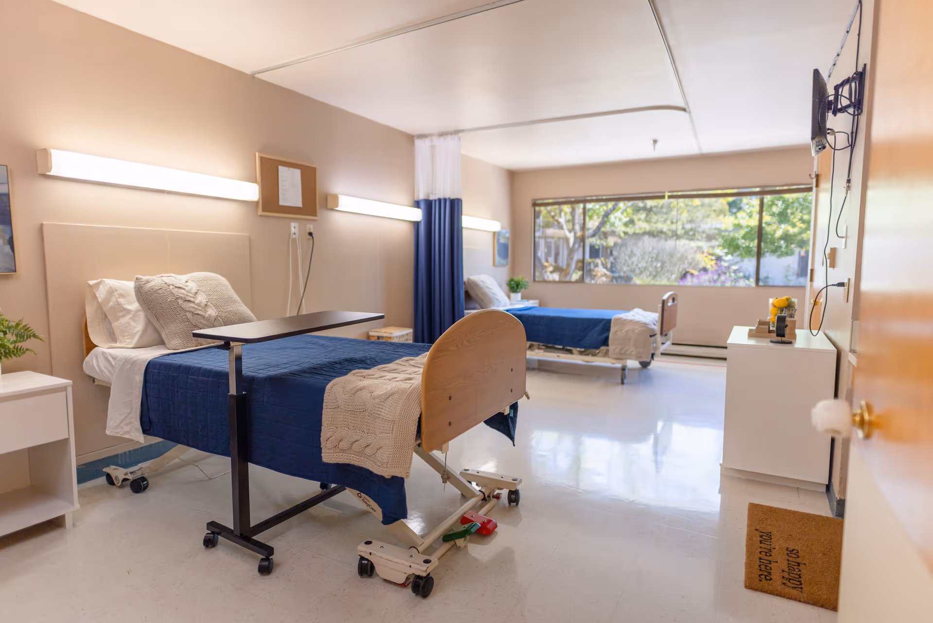 A bright and clean nursing facility bedroom with two hospital beds, each made with blue blankets and beige knitted throws. There is a large window at the far end letting in natural light, a small white nightstand with a plant, a wall-mounted TV, and a white cabinet. The room has light-colored walls and flooring, with a welcome mat on the floor near the door.