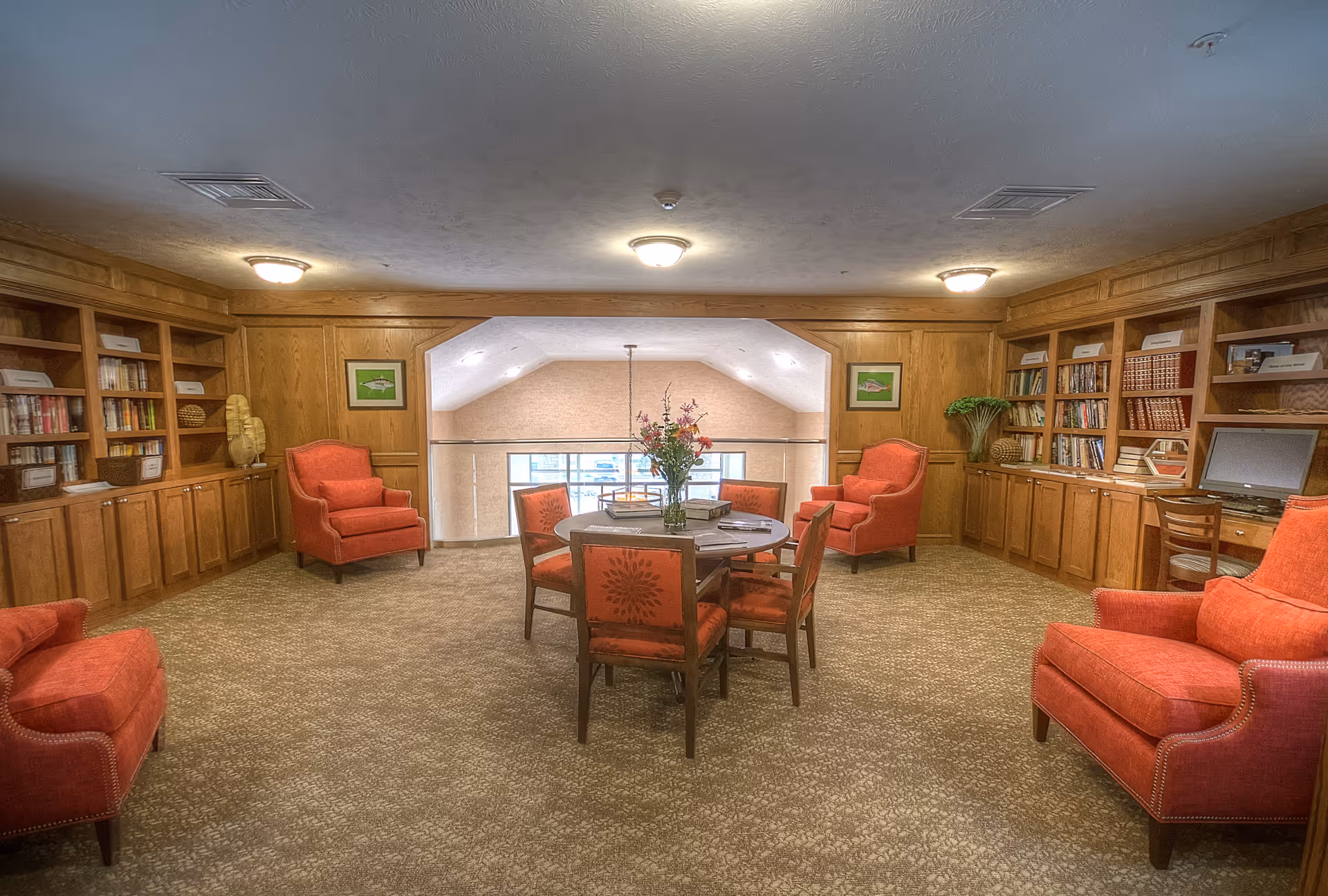 A cozy common area with beige carpet and wood-paneled walls featuring built-in bookshelves filled with books and decorative items. The room has four red upholstered armchairs arranged around a round wooden table with six matching chairs. A vase with flowers sits on the table. Two framed pictures hang on the walls, and a computer workstation is visible on the right side. The ceiling has recessed lighting fixtures.
