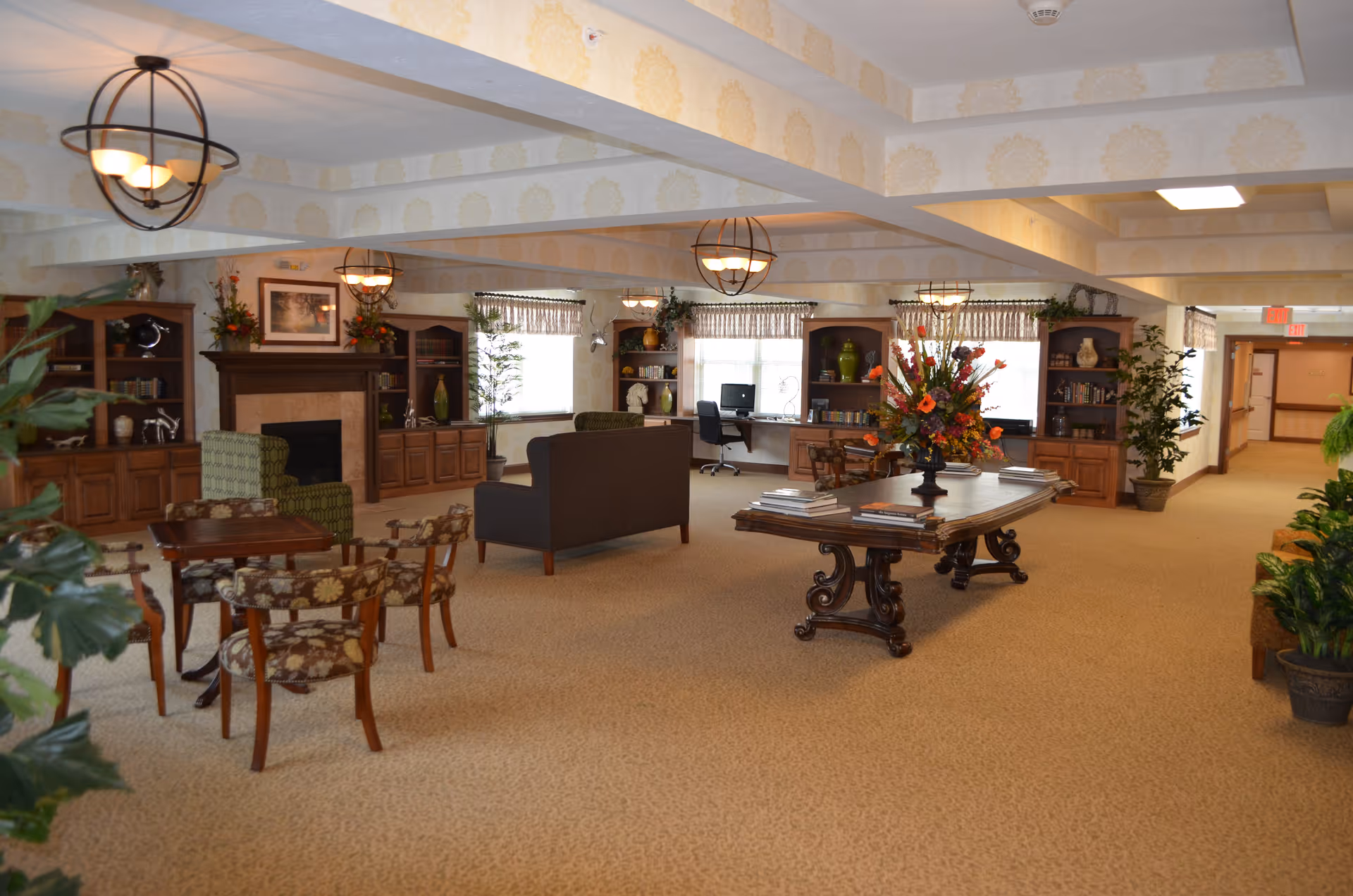 Spacious senior living common room with seating areas, a fireplace, tables, bookshelves, and decorative plants.