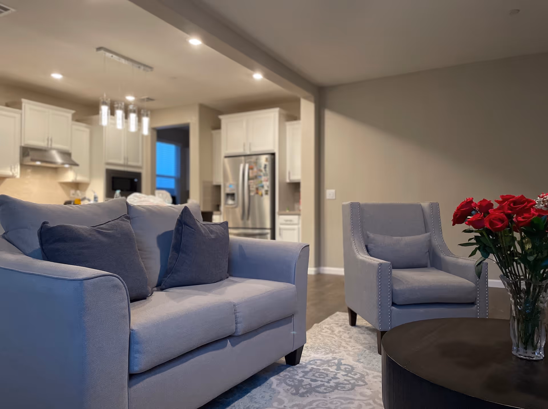 A cozy living room area with a light gray sofa and matching armchair, both with dark gray cushions. A round black coffee table with a vase of red roses is in front of the armchair. In the background, there is a modern kitchen with white cabinets, stainless steel appliances, and pendant lighting.