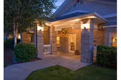 Entrance to a residential building at dusk with warm lighting, brick pillars, a covered porch, and surrounding greenery including bushes and a tree.