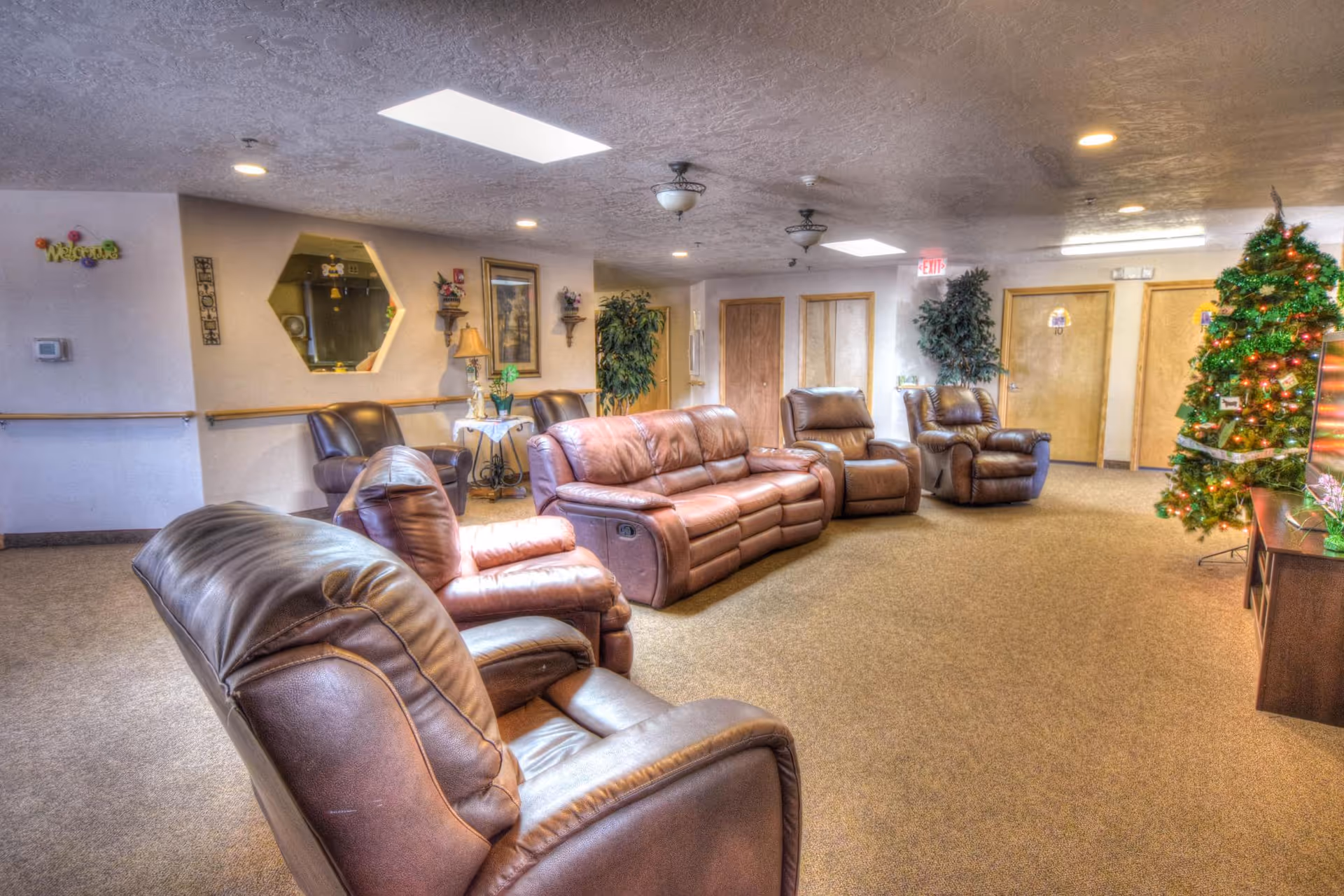 A cozy living room area in a senior living facility featuring several brown leather recliners and a matching sofa arranged around a TV stand. The room is decorated with a Christmas tree adorned with lights and ornaments. The walls have framed artwork, a hexagonal mirror, and some small shelves with decorative items. There are ceiling lights and fans, and the carpet is beige. Doors and plants are visible in the background.