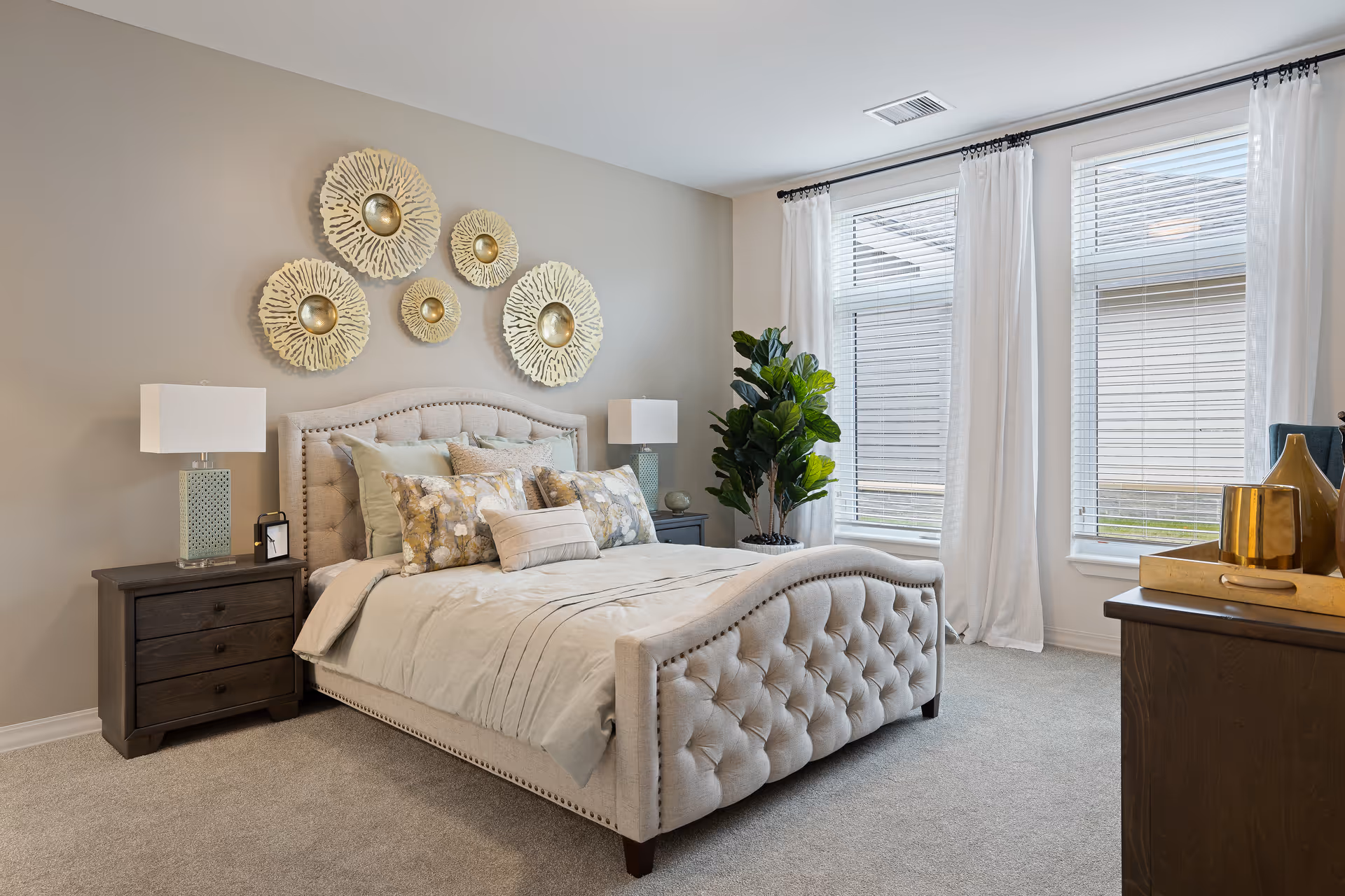 A well-lit bedroom featuring a tufted beige upholstered bed with multiple decorative pillows. On either side of the bed are dark wooden nightstands with modern lamps. Above the bed, there are five decorative circular wall hangings with a gold center. Large windows with white curtains allow natural light to fill the room. A green potted plant is placed near the windows, and a dark wooden dresser with gold decorative items is visible on the right side.