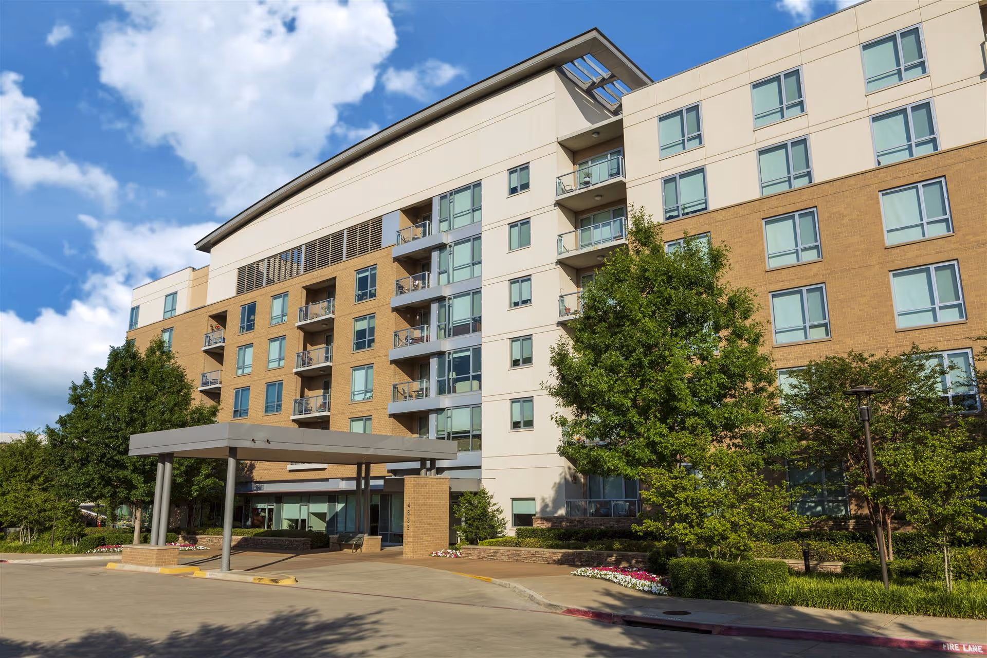 Exterior view of a multi-story senior living facility building with balconies, large windows, and a covered entrance. The building is surrounded by trees, shrubs, and a well-maintained garden area under a partly cloudy blue sky.