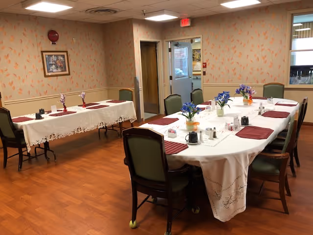 Dining room with two long tables set with white tablecloths, burgundy placemats, floral centerpieces and chairs on a wood floor.
