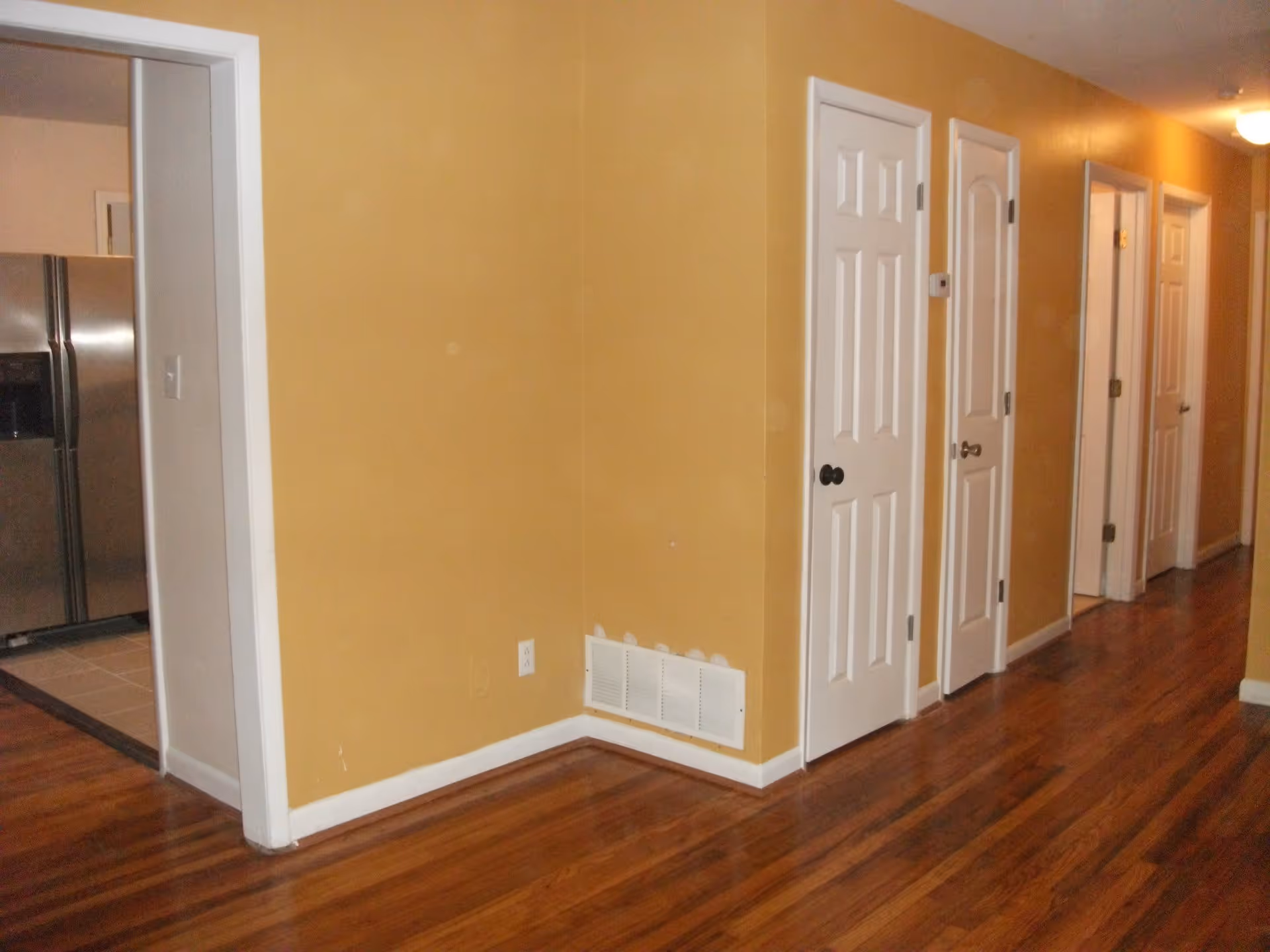 Interior hallway with wooden floor and mustard yellow walls. Several white doors line the hallway on the right side. On the left, there is an open doorway leading to a kitchen area with a stainless steel refrigerator.