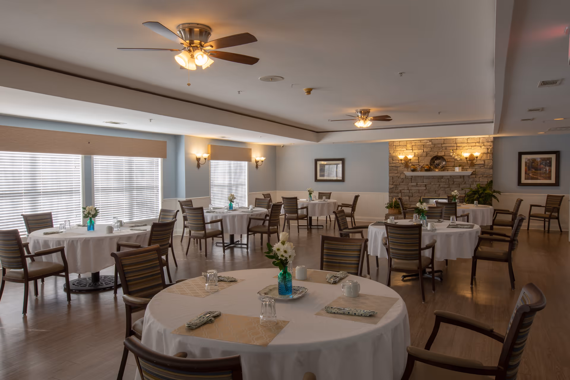 A dining room with multiple round tables covered with white tablecloths, each set with napkins, glasses, and small flower vases. The room has wooden chairs with striped cushions, ceiling fans with lights, large windows with blinds, and a stone fireplace with wall sconces.