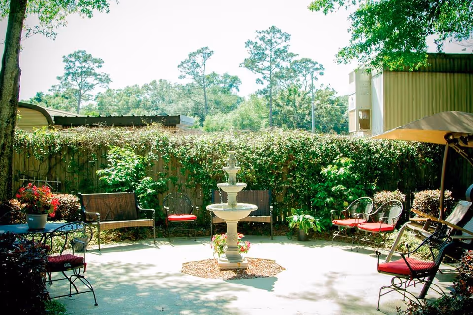 Outdoor patio area with a central three-tiered water fountain surrounded by various metal chairs and benches with red cushions. The space is enclosed by a wooden fence covered in green vines and plants, with trees visible in the background under a bright sky.