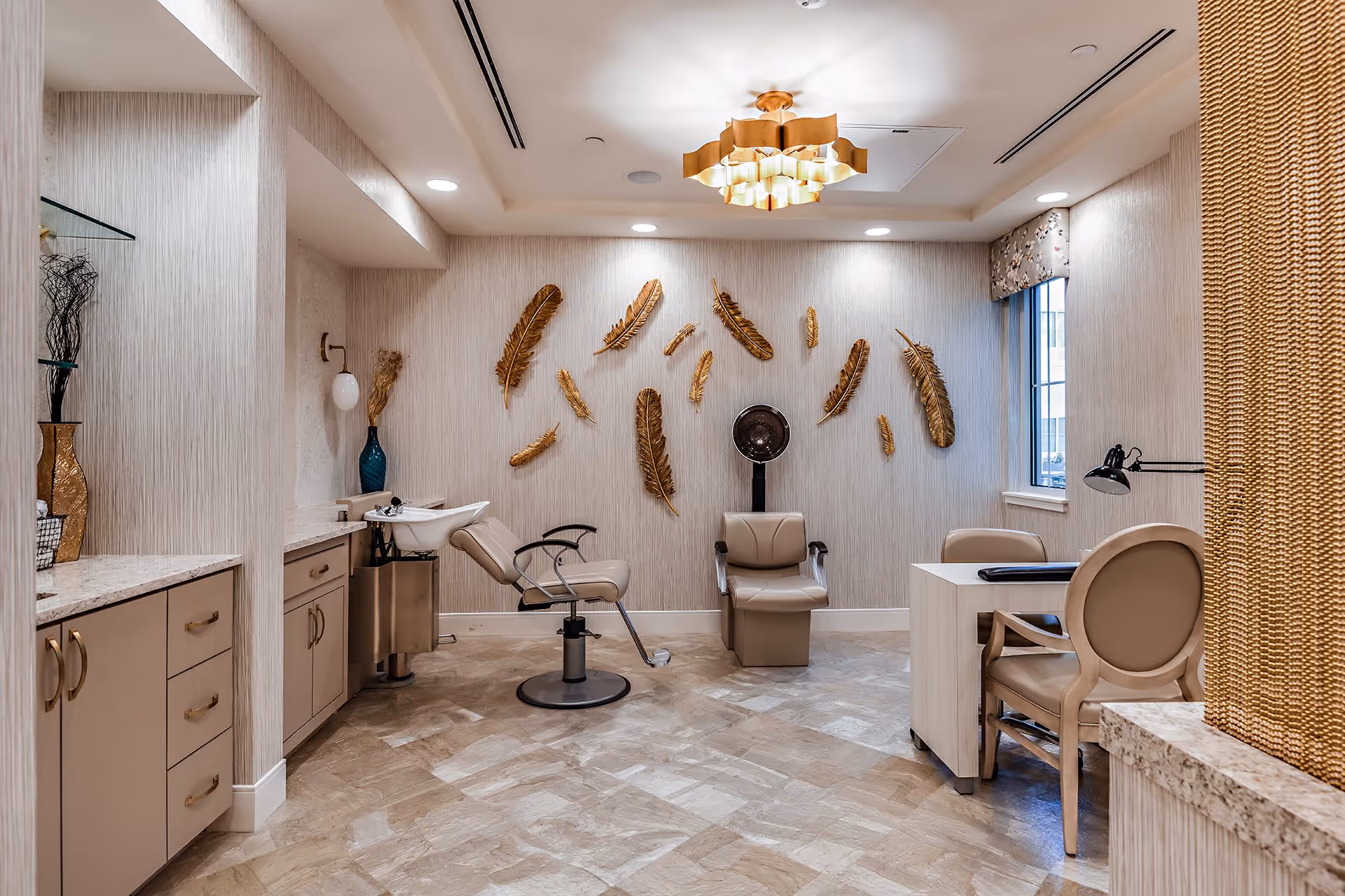Bright salon-style room with a styling chair and shampoo sink, a manicure table and chairs, and decorative gold feather wall art on the back wall.
