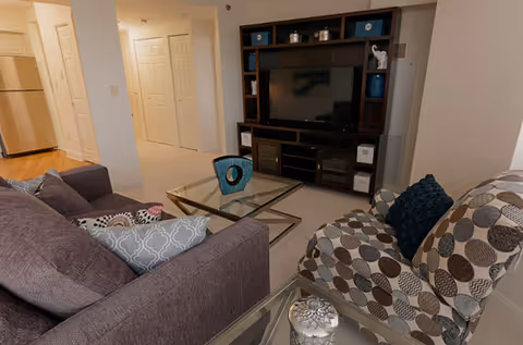 A cozy living room area with a dark gray sofa adorned with patterned pillows, a patterned armchair with a dark blue pillow, a glass coffee table with a decorative blue vase, and a dark wooden entertainment center with a flat-screen TV and decorative items. The room has light-colored walls and carpet, with a view into a hallway and kitchen area in the background.