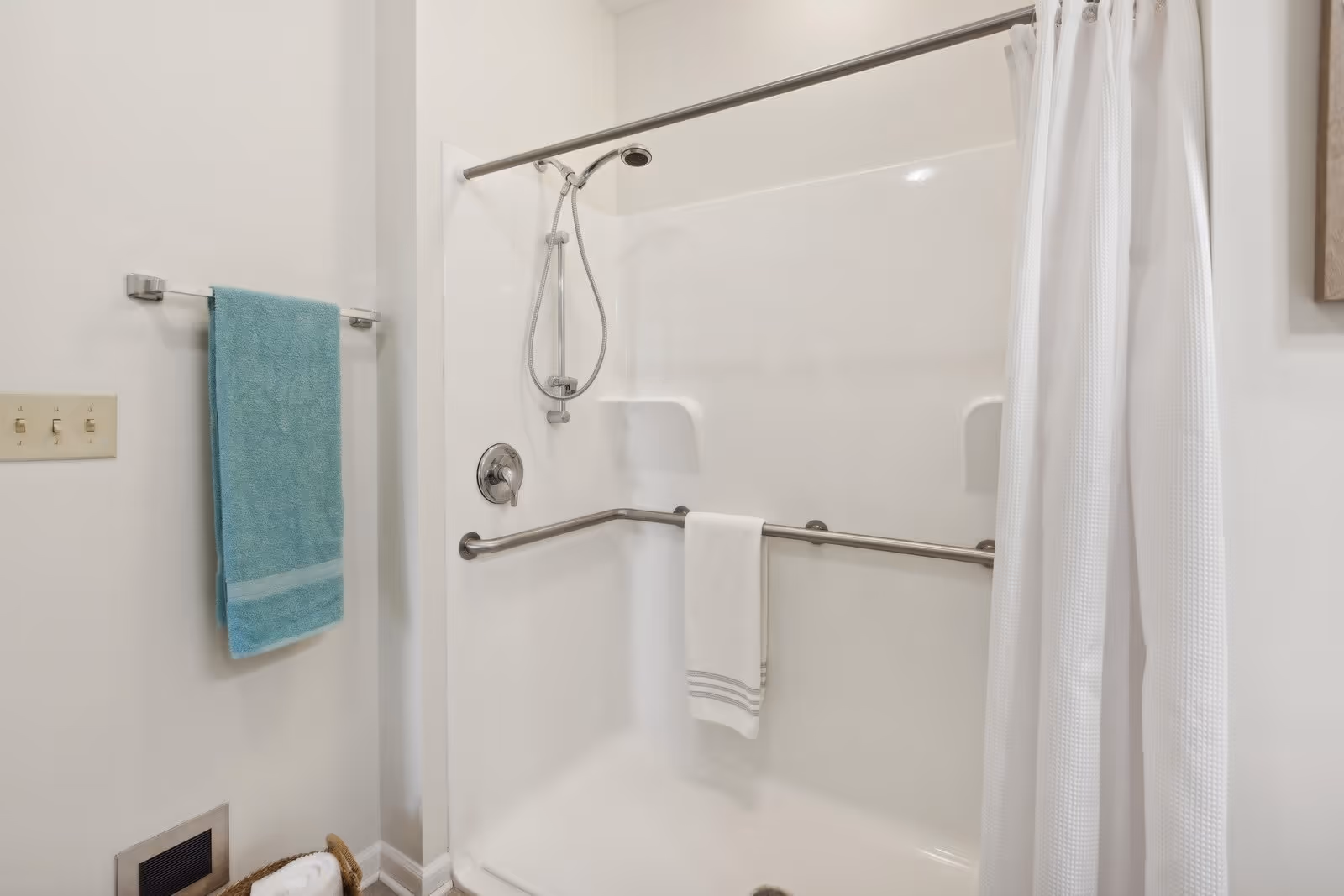 A clean bathroom shower area with a white shower curtain partially open, a handheld showerhead, and a horizontal grab bar with a white towel hanging on it. A blue towel hangs on a towel rack on the adjacent wall, and a basket with rolled towels is visible on the floor.