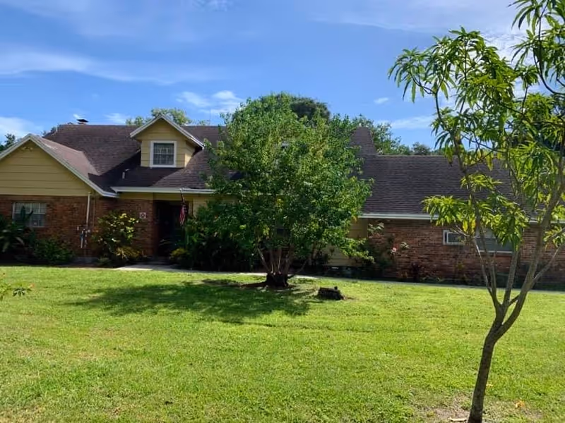 A single-story brick and yellow siding house with a dark shingled roof, surrounded by a green lawn and trees under a blue sky with some clouds.