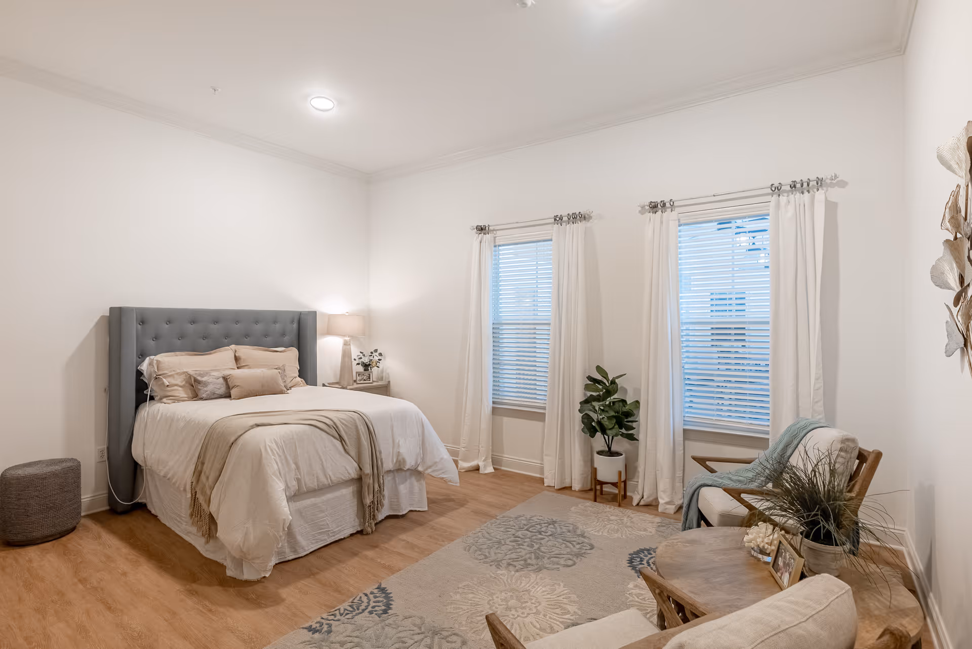 A cozy bedroom with a large bed featuring a tufted gray headboard, beige bedding, and multiple pillows. There is a bedside table with a lamp and a small plant. The room has two windows with white curtains and blinds, a patterned area rug, and a seating area with two chairs and a wooden coffee table decorated with plants and picture frames. The walls are white and the floor is wooden.