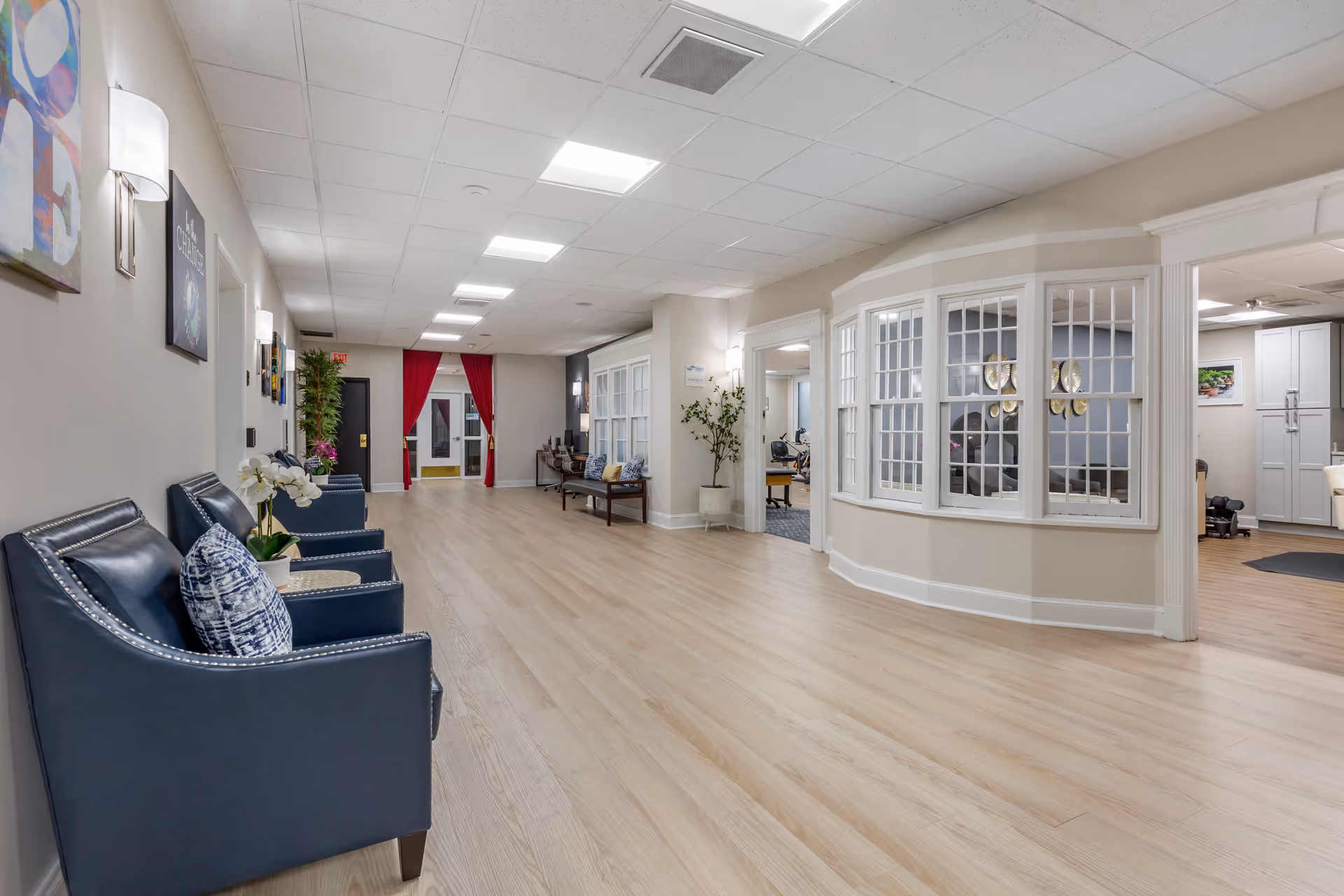 A spacious, well-lit hallway in Thomas House Retirement Community with light wood flooring and beige walls. On the left side, there are several dark blue armchairs with patterned pillows and small tables with plants. The hallway leads to a door with red curtains. On the right side, there is a large windowed room with white trim and a glimpse of a kitchen area with white cabinets and appliances. The hallway is decorated with wall art and plants.
