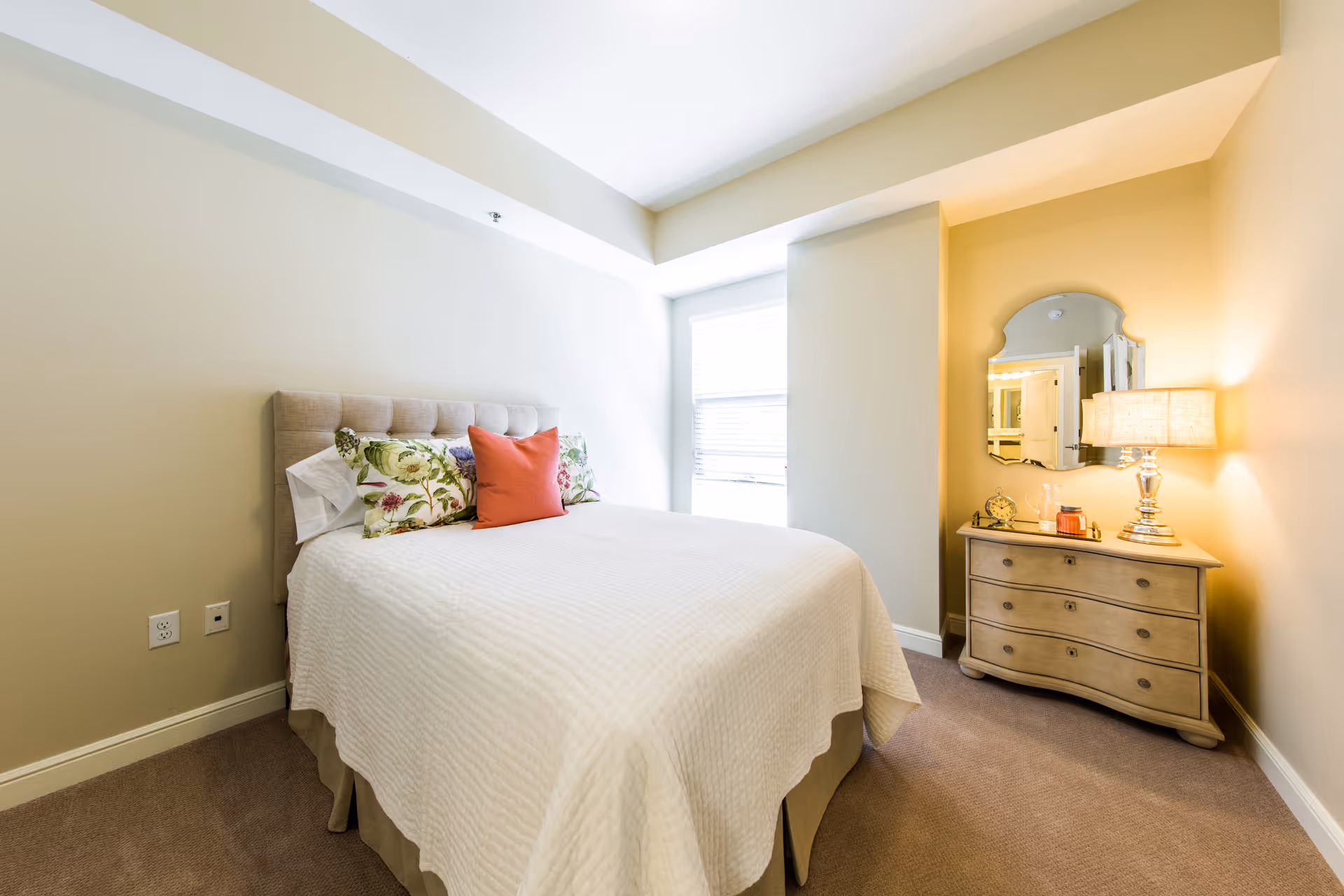 A bright bedroom with a neatly made bed featuring a white quilt and decorative pillows, including floral and orange ones. To the right of the bed is a wooden dresser with a curved front, topped with a lamp, a decorative clock, and other small items. A mirror hangs above the dresser, reflecting part of the room. The walls are painted in light beige tones, and there is a window with blinds letting in natural light.
