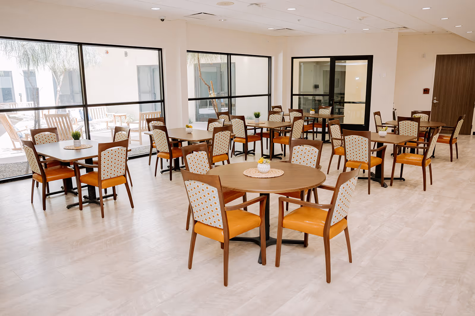 A bright and spacious dining room with multiple wooden tables and chairs arranged neatly. The chairs have orange seats and patterned backs. Large windows let in natural light and provide a view of an outdoor patio area with wooden benches and plants.