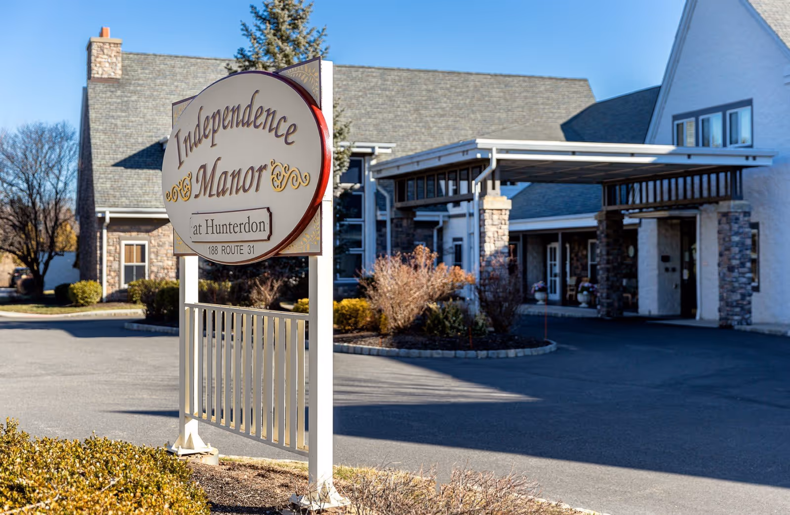 Exterior view of Independence Manor at Hunterdon showing the facility's sign in the foreground and the building entrance with a covered drop-off area in the background under a clear blue sky.