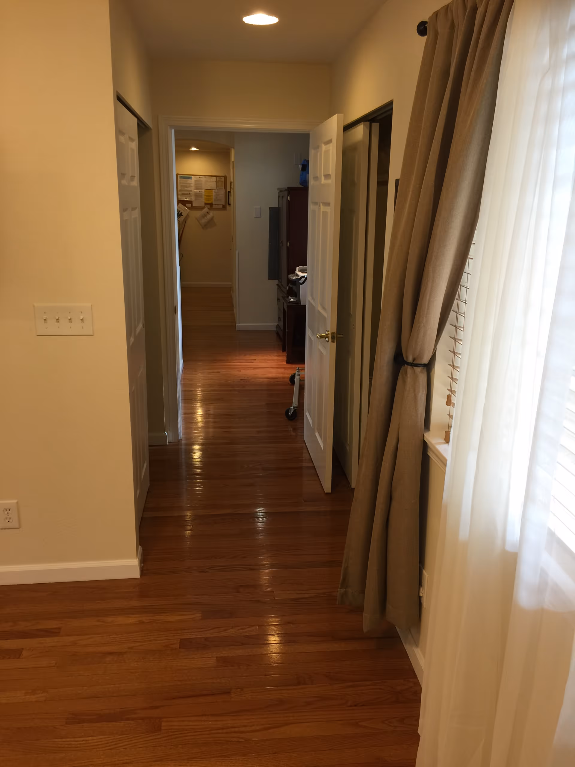 A hallway with polished wooden floors leading to an open door. On the right side, there is a window with beige curtains and white sheer drapes. The walls are painted light beige, and there is a light fixture on the ceiling illuminating the hallway.
