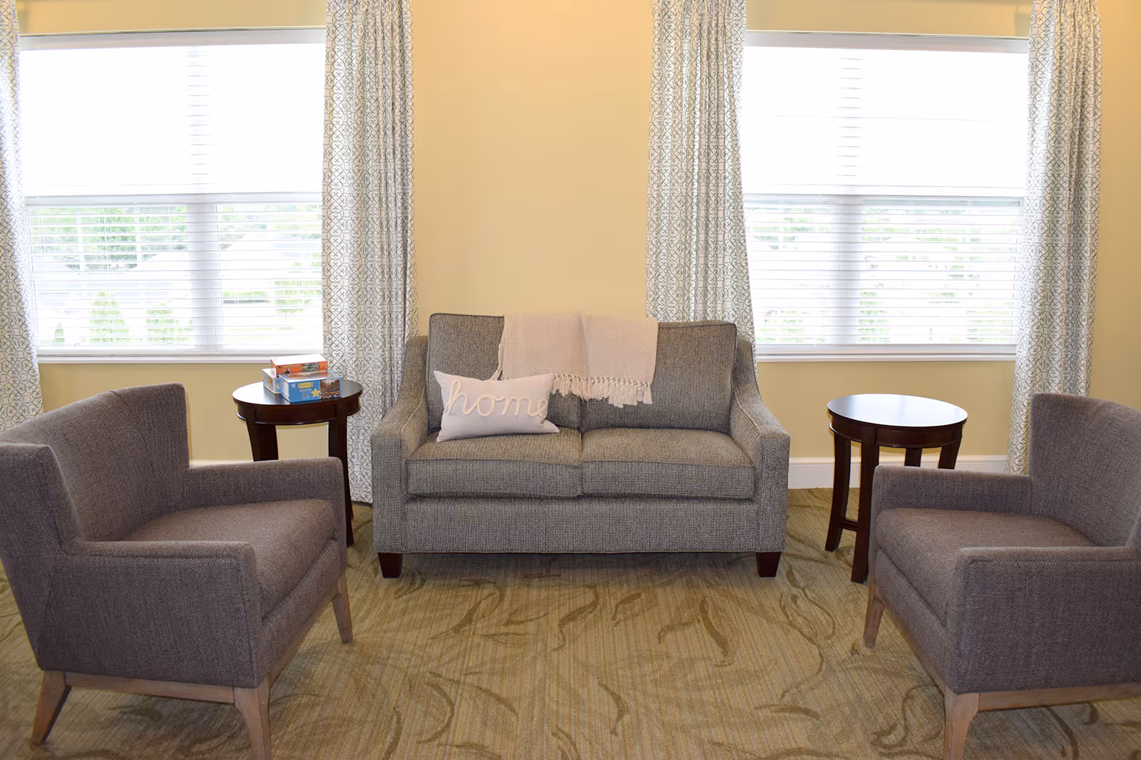 A cozy seating area in a living room with a gray loveseat adorned with a light pink throw blanket and a pillow that says 'home'. Two matching gray armchairs face the loveseat, and there are two small round wooden side tables next to the chairs. The room has large windows with patterned curtains and light yellow walls.
