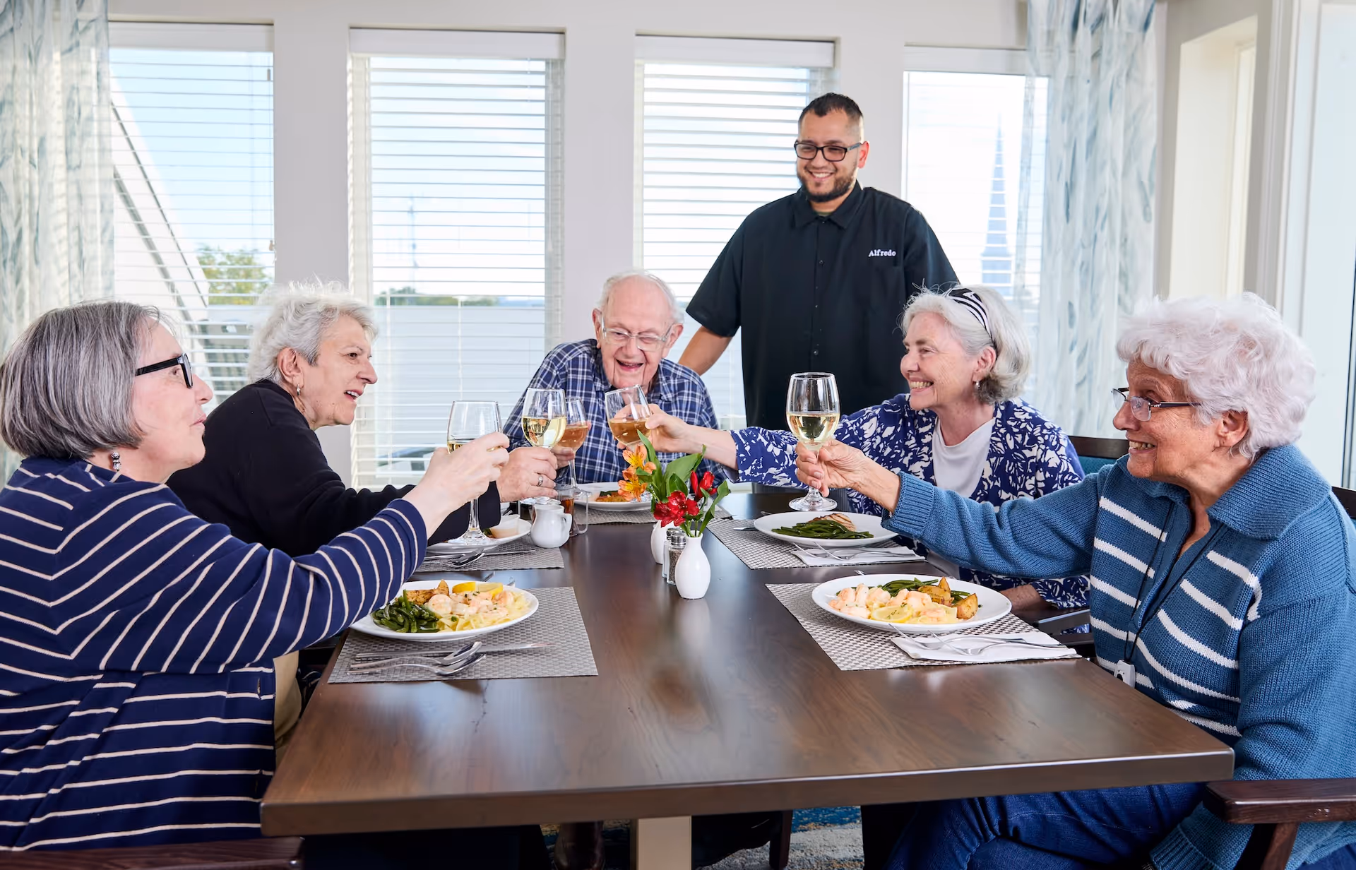 Five elderly people sitting around a dining table raising their glasses in a toast, with a smiling staff member standing behind them. Plates of food and glasses of white wine are on the table, and large windows with blinds are in the background.