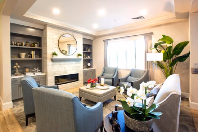 A cozy living room with a light gray sofa, two blue armchairs, and two gray chairs arranged around a square ottoman. The room features a modern fireplace with a round mirror above it, built-in shelves with decorative items, a large window with curtains, a floor lamp, and a potted plant. A small table with white orchids is in the foreground.