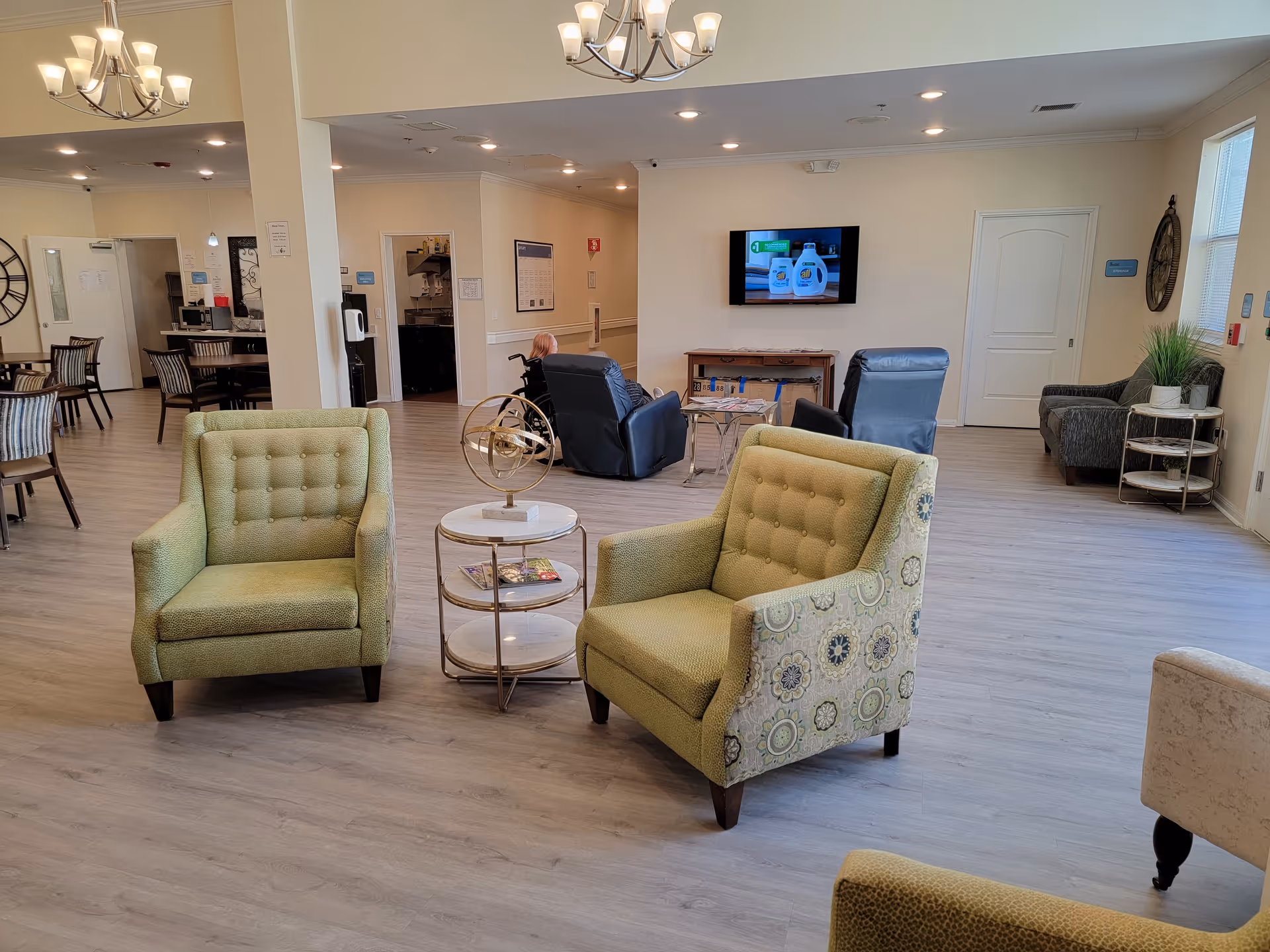 A spacious and well-lit common area in a senior living facility with light wood flooring, two green patterned armchairs facing each other with a small round table between them, additional seating including recliners and chairs, a wall-mounted TV, and a chandelier overhead.