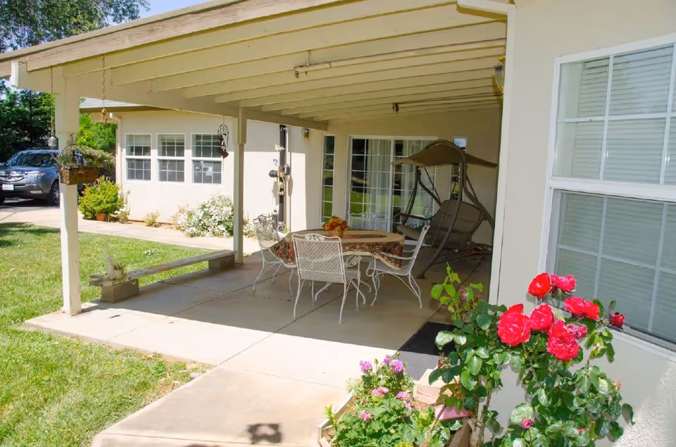 Covered outdoor patio with a dining table and chairs, a swing, and potted flowers beside a light-colored building.