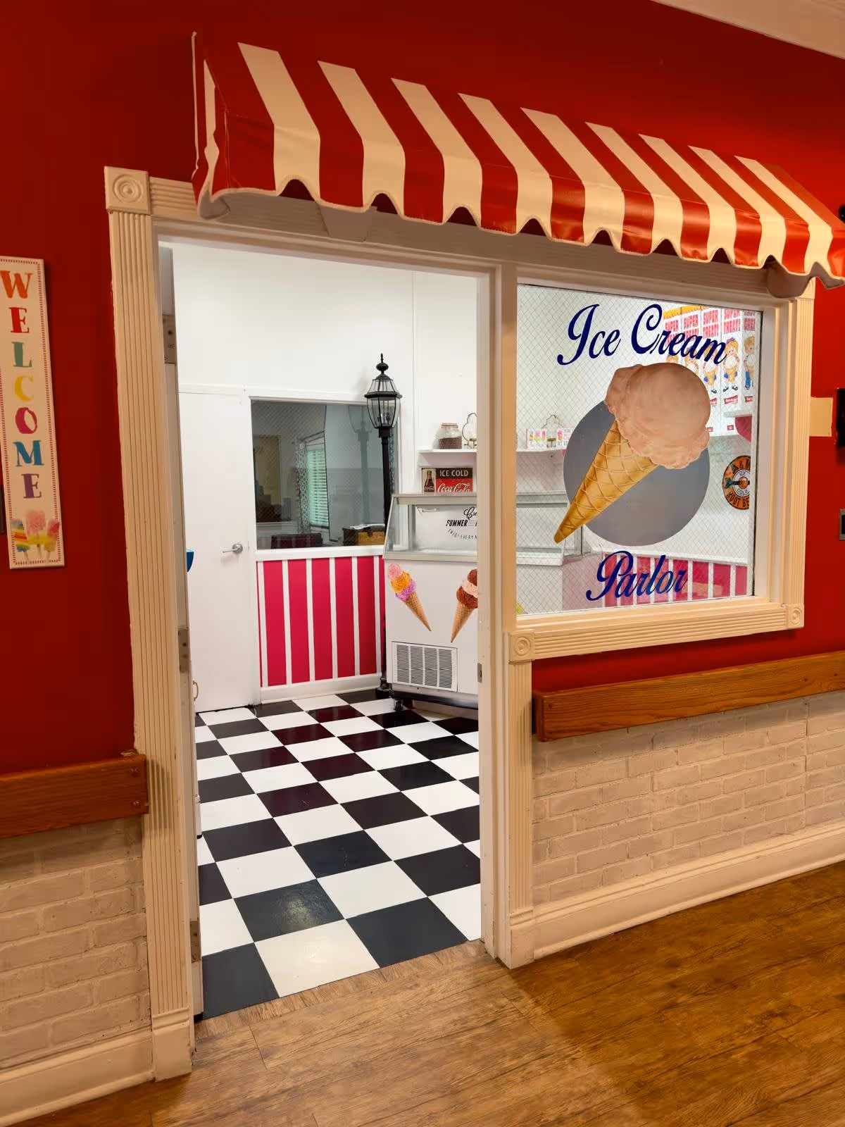 Entrance to a vintage-style indoor ice cream parlor with a red-and-white striped awning, checkerboard floor, and ice cream cone signage.