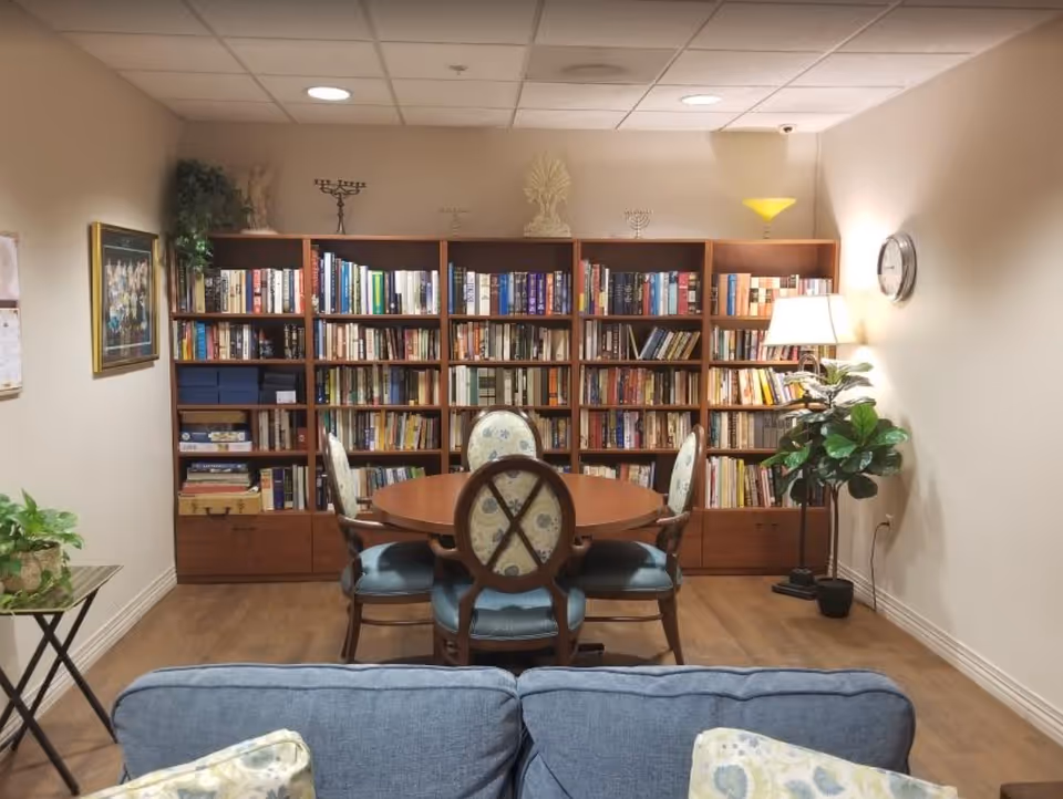 Cozy interior sitting area with a round table and upholstered chairs in front of a large bookshelf, a couch in the foreground and a floor lamp to the right.
