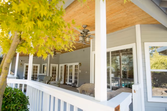 Covered outdoor porch area of a senior living facility with white railing, ceiling fans, wicker chairs, and multiple glass doors and windows, surrounded by greenery.