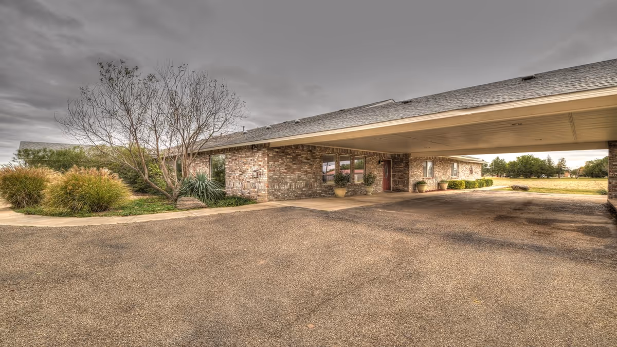 Front entrance with a covered drive (porte-cochère) of a single-story brick building surrounded by shrubs and open grounds.