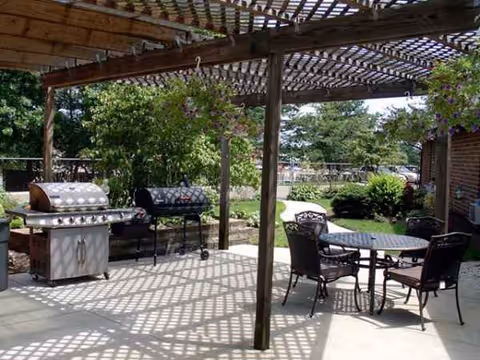 Covered outdoor patio with grill, table and chairs, and garden greenery.