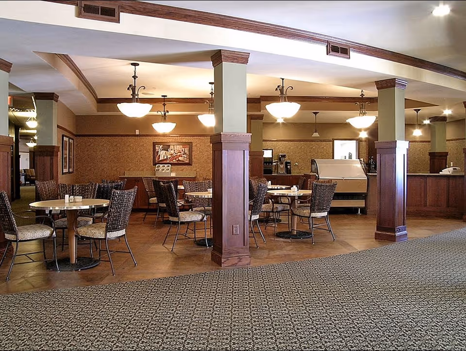 Interior view of a dining area in a senior living facility with round tables and wicker chairs. The room features wooden columns, warm lighting fixtures hanging from the ceiling, and a carpeted floor with a patterned design. There is a counter area in the background with coffee machines and other equipment.