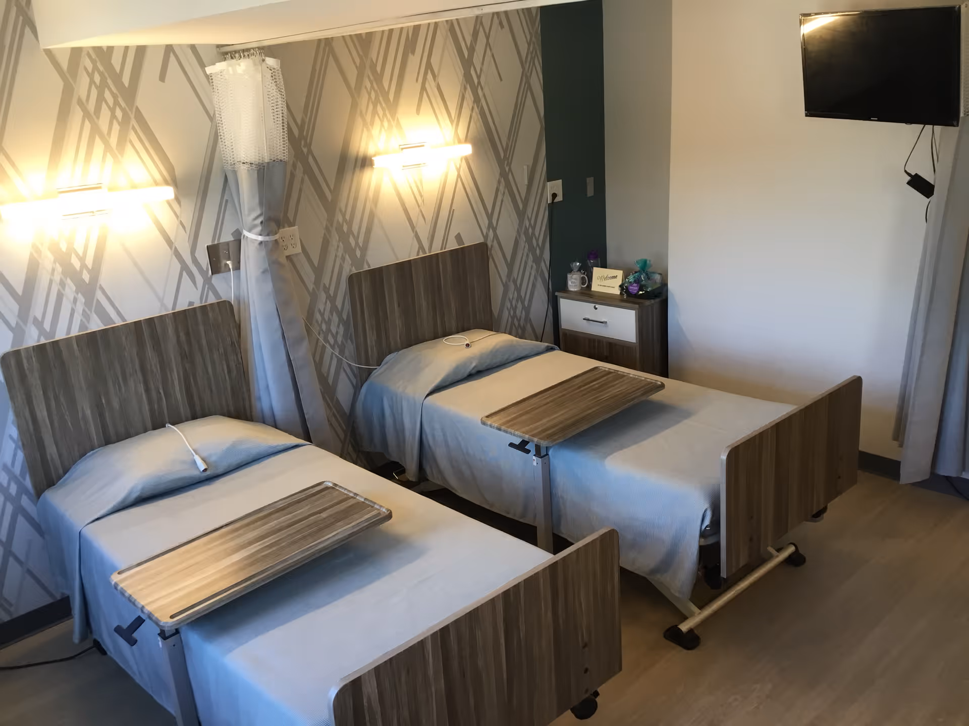 A room with two hospital-style beds, each with a wooden headboard and footboard, light blue bedding, and an adjustable overbed table. The wall behind the beds has a patterned wallpaper with two wall-mounted lights. There is a small nightstand between the beds with a welcome sign and some decorative items. A flat-screen TV is mounted on the wall to the right.