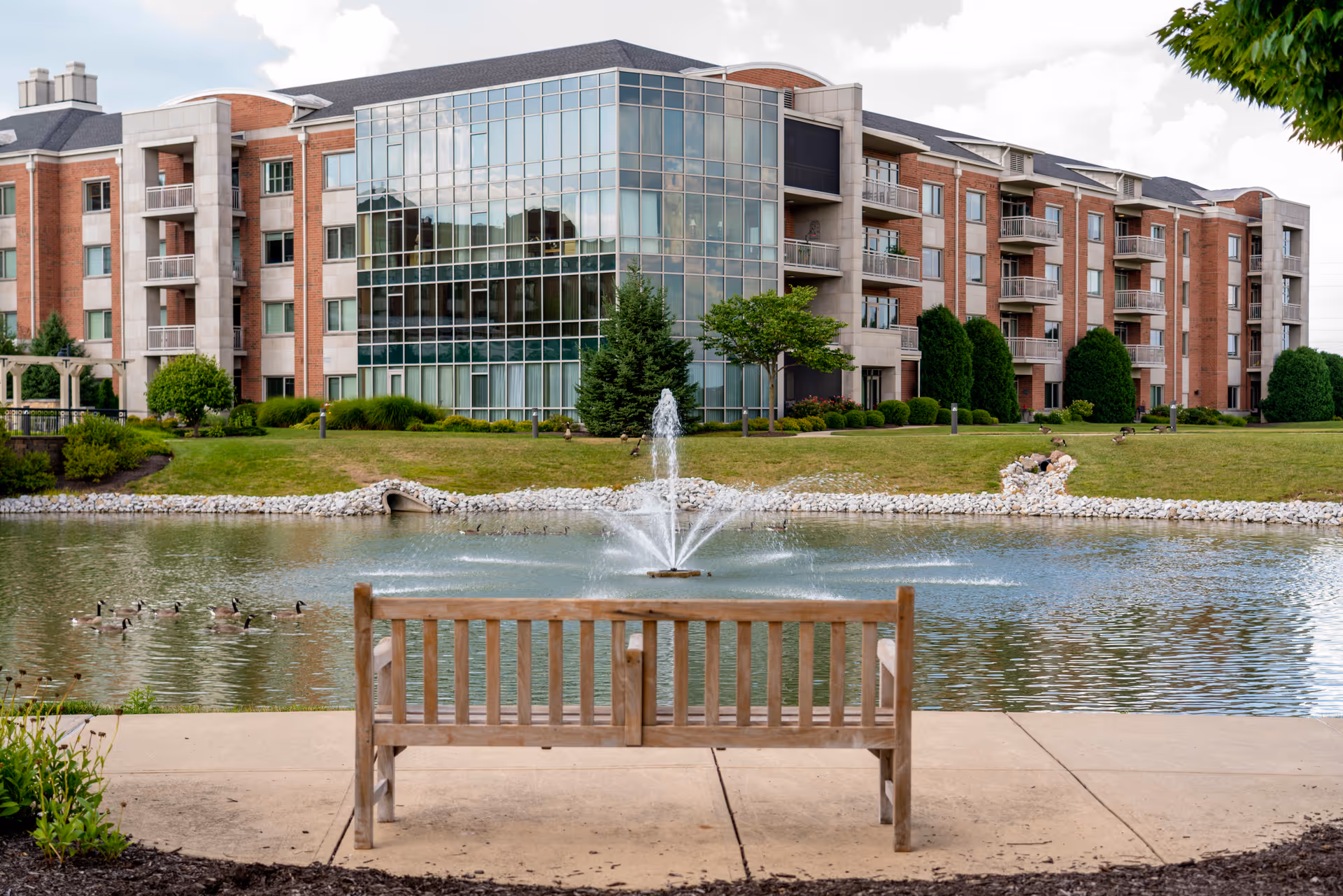 A wooden bench facing a pond with a water fountain in the center, surrounded by green grass and trees. In the background, there is a large multi-story brick building with many windows and balconies.