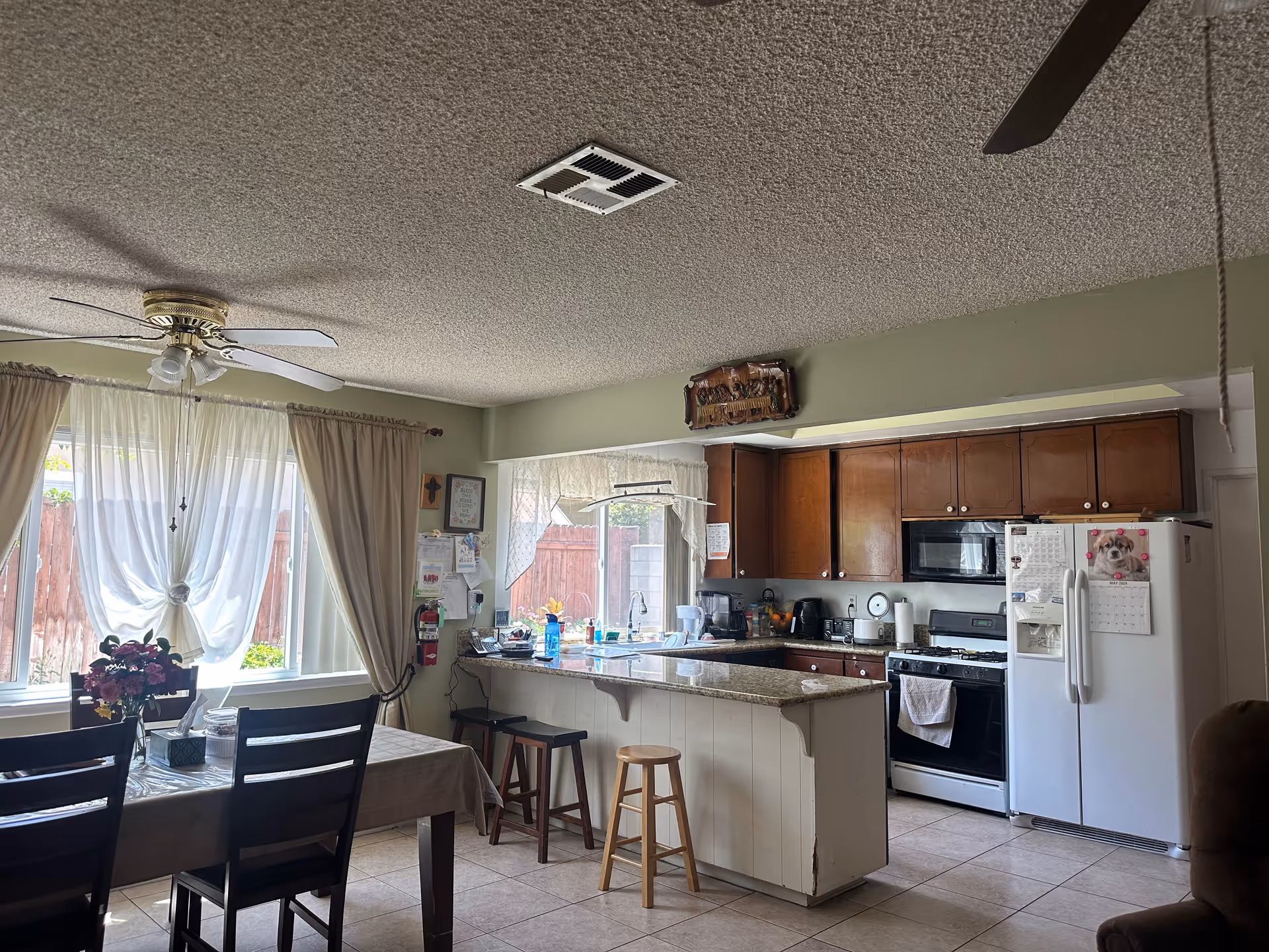 Interior view of a kitchen and dining area with a table and chairs near a window with curtains. The kitchen has wooden cabinets, a white refrigerator, a stove, a microwave, and a granite countertop island with stools. There is a ceiling fan above the dining table and various kitchen appliances on the counters.