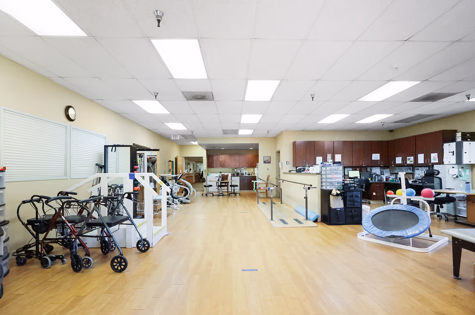 A spacious rehabilitation or physical therapy room with wooden flooring, parallel bars for walking exercises, various exercise equipment including a small trampoline, walkers, and chairs. The room has a drop ceiling with fluorescent lights and a workstation with computers and office supplies on the right side.