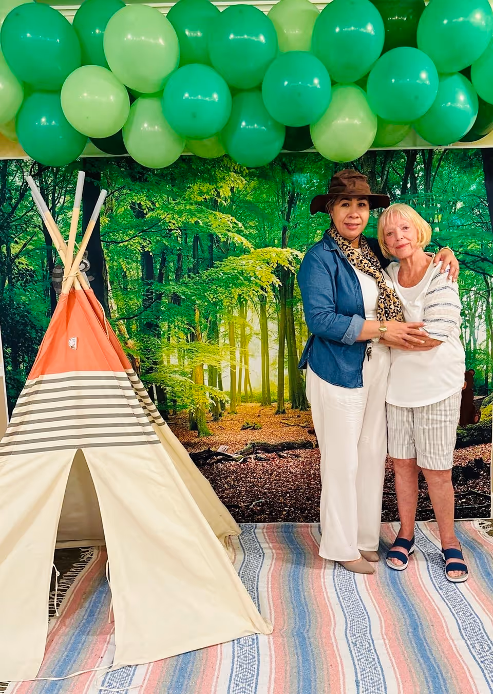 Two women standing and hugging indoors in front of a wall mural depicting a green forest. To the left is a small beige and orange striped teepee tent, and above them are green balloons attached to the ceiling. The floor is covered with a striped rug.