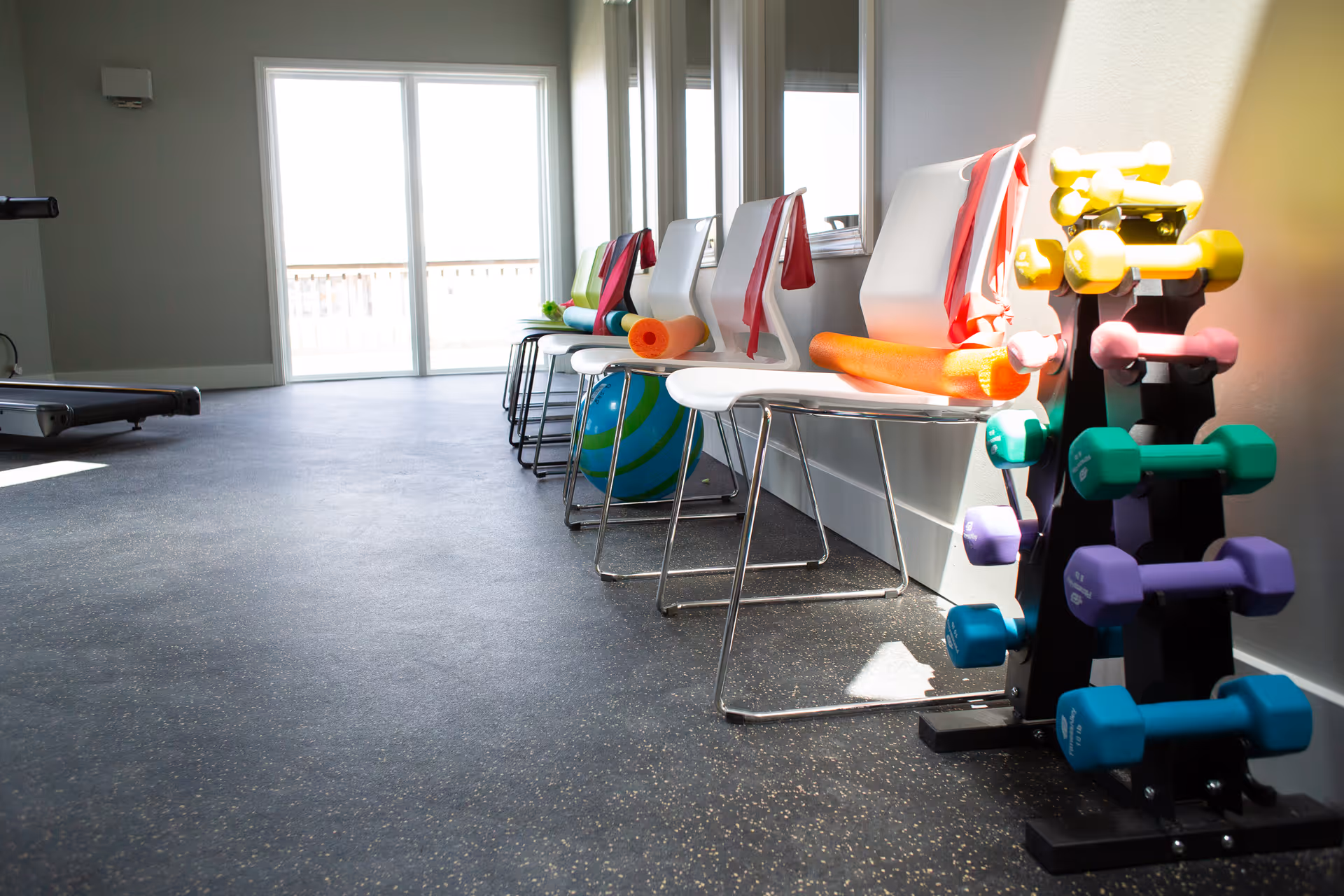 A bright exercise room with a row of white chairs along the wall, each with a colorful resistance band and foam roller placed on them. Next to the chairs is a rack holding various colorful dumbbells. Large windows and a glass door allow natural light to fill the room.