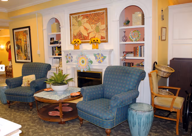 A cozy living room area with two blue checkered armchairs, a round wooden coffee table with a plant and decorative plates, a fireplace decorated with paper flowers and sunflowers, built-in shelves with books and decorative items, and a wooden chair with a basket on the side.