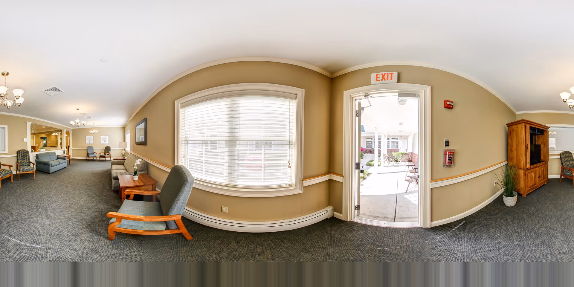 Bright communal lounge with seating, tables, a large window, and an open EXIT door leading to an outdoor courtyard.