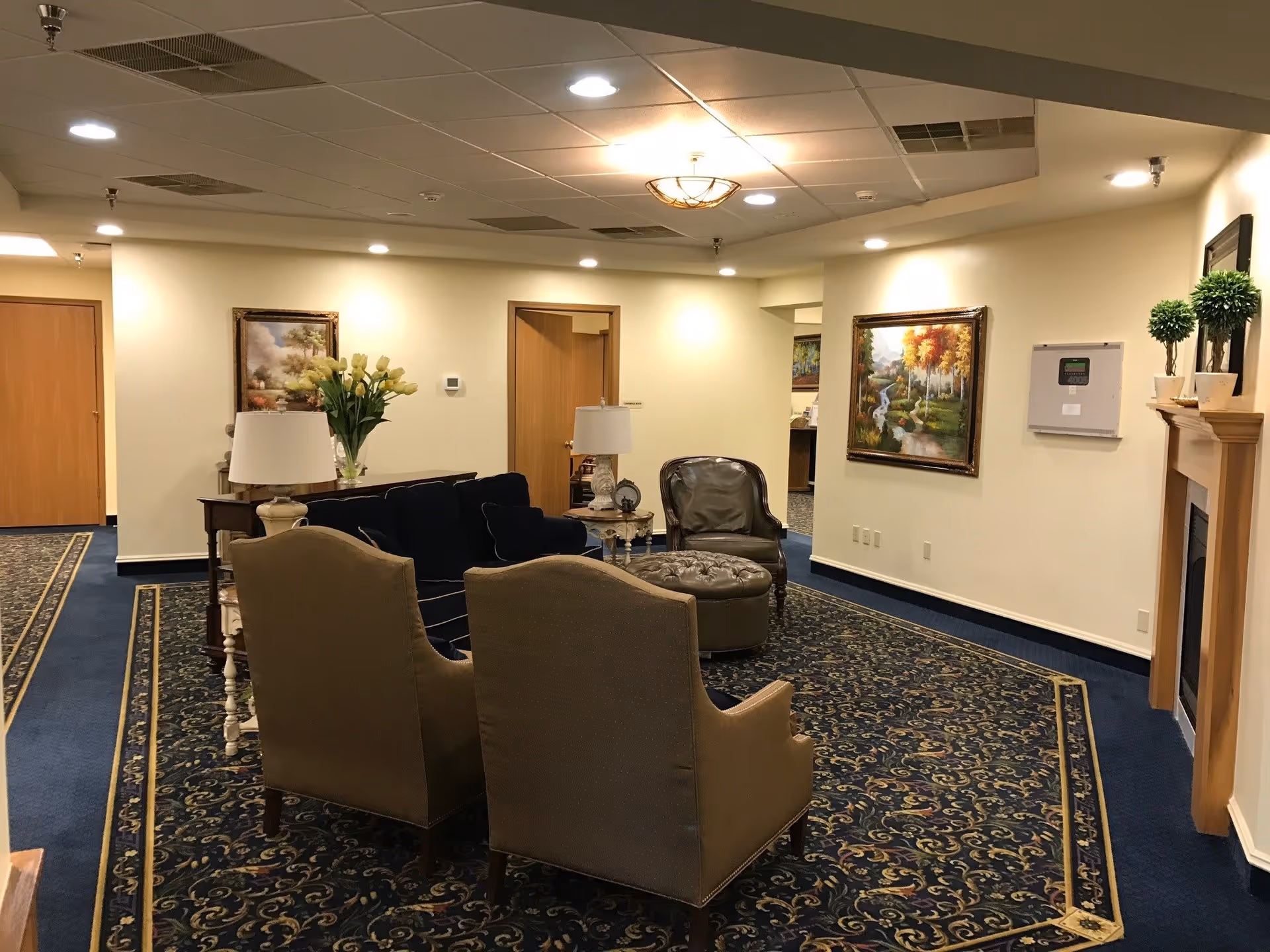 A cozy sitting area in a senior living facility with a dark blue couch, two beige armchairs, a round tufted ottoman, and two side tables with lamps. The room has a patterned carpet with blue and gold designs, cream-colored walls, framed landscape paintings, and a wooden fireplace mantel decorated with small potted plants. There are several doors and a hallway visible in the background.