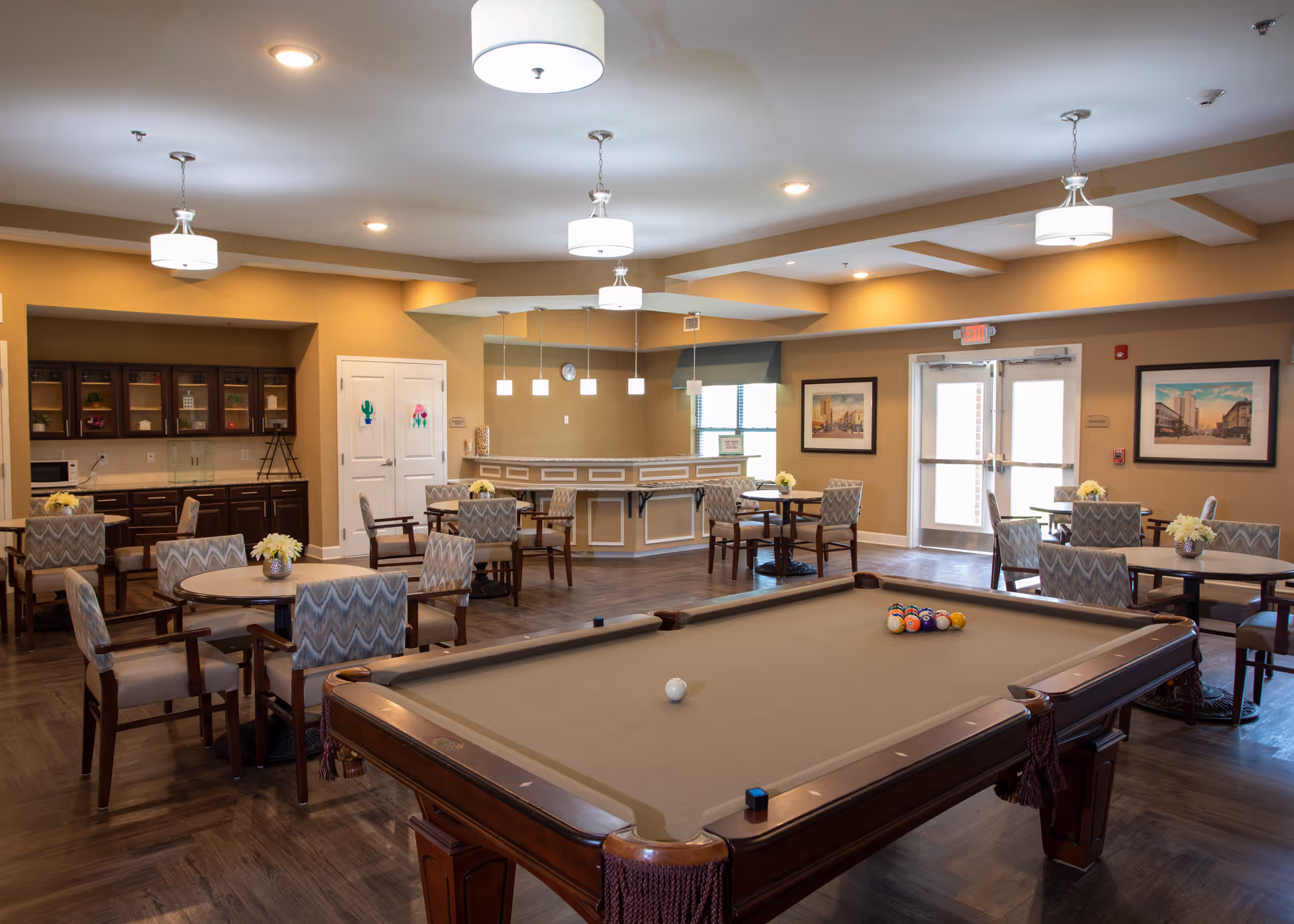 Spacious senior living common room with a pool table in the foreground, round tables and chairs, and a kitchenette in the background.