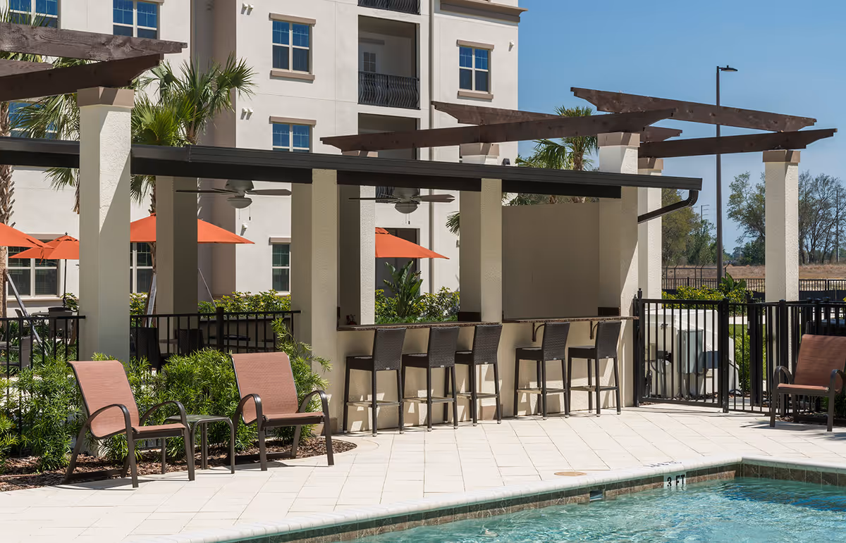 Outdoor pool area with lounge chairs, bar seating under pergolas and a multi-story building in the background.