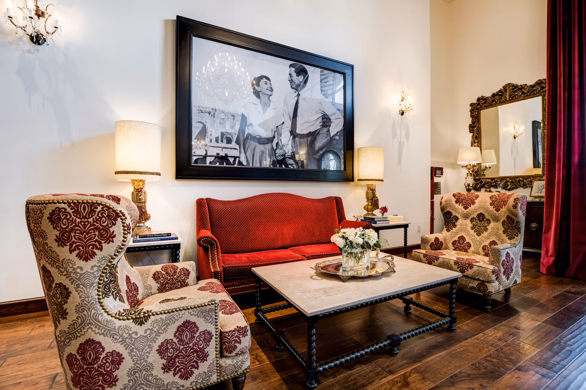 A cozy living room area with a red upholstered sofa and two patterned armchairs around a rectangular coffee table. The table has a silver tray with a vase of white flowers. Two table lamps with beige shades are on either side of the sofa. A large black and white framed photograph of a smiling couple hangs on the wall above the sofa. There is a large ornate mirror on the adjacent wall and red curtains partially visible on the right side. The room has wooden flooring and soft lighting.
