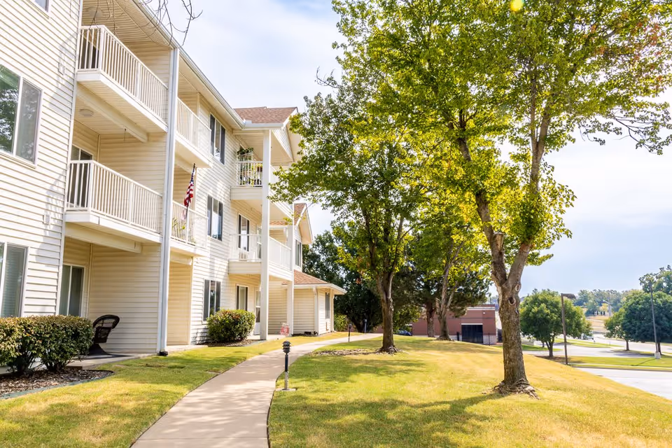 Exterior view of a multi-story residential building with balconies, a paved walkway, green grass, and several trees under a partly cloudy sky.