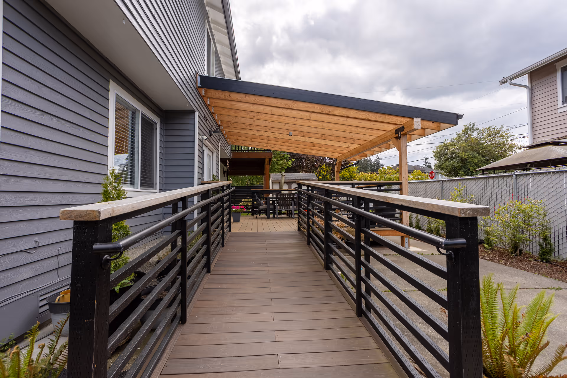 Outdoor wooden ramp with black metal railings leading to a covered patio area attached to a gray building. The patio has a wooden pergola roof and outdoor seating. There are plants and a fence along the side of the ramp.